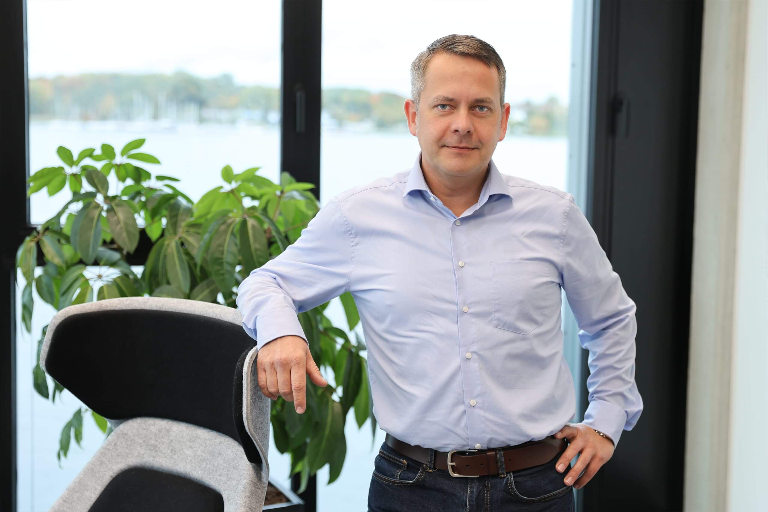 Ronny Schröder Ronny Schröder steht an einen Stuhl lehnend in seinem Büro vor großen Fenstern.
